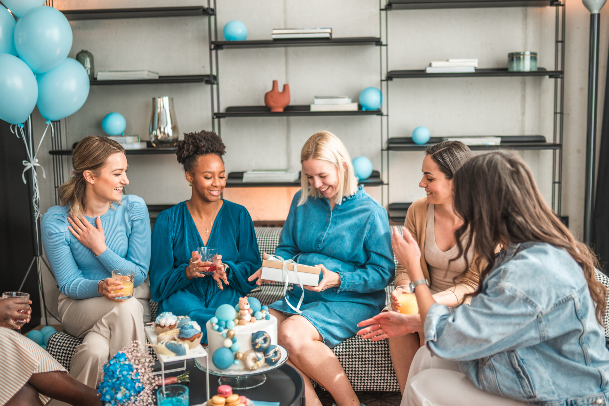 In a room filled with baby shower decor, a small group of diverse female friends gathers to celebrate their pregnant blonde friend. They gift her with love and presents while enjoying each other's company.